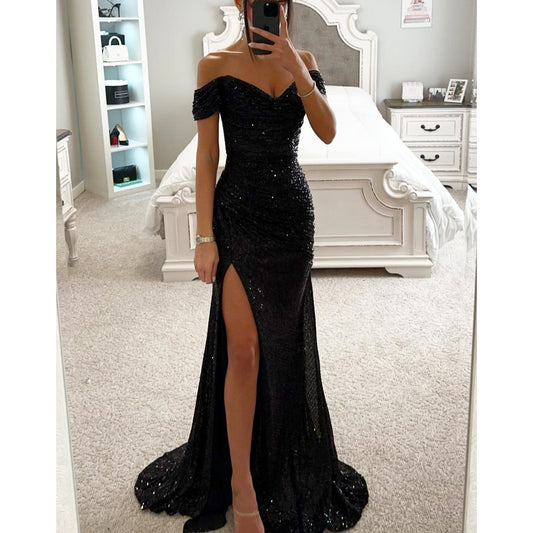 Person wearing a black off-shoulder evening gown with a high slit in a bedroom.