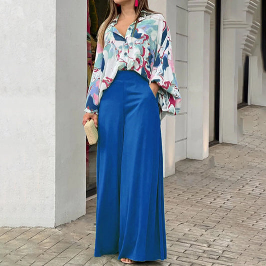 Woman wearing a colorful floral blouse and blue pants standing against a white wall.